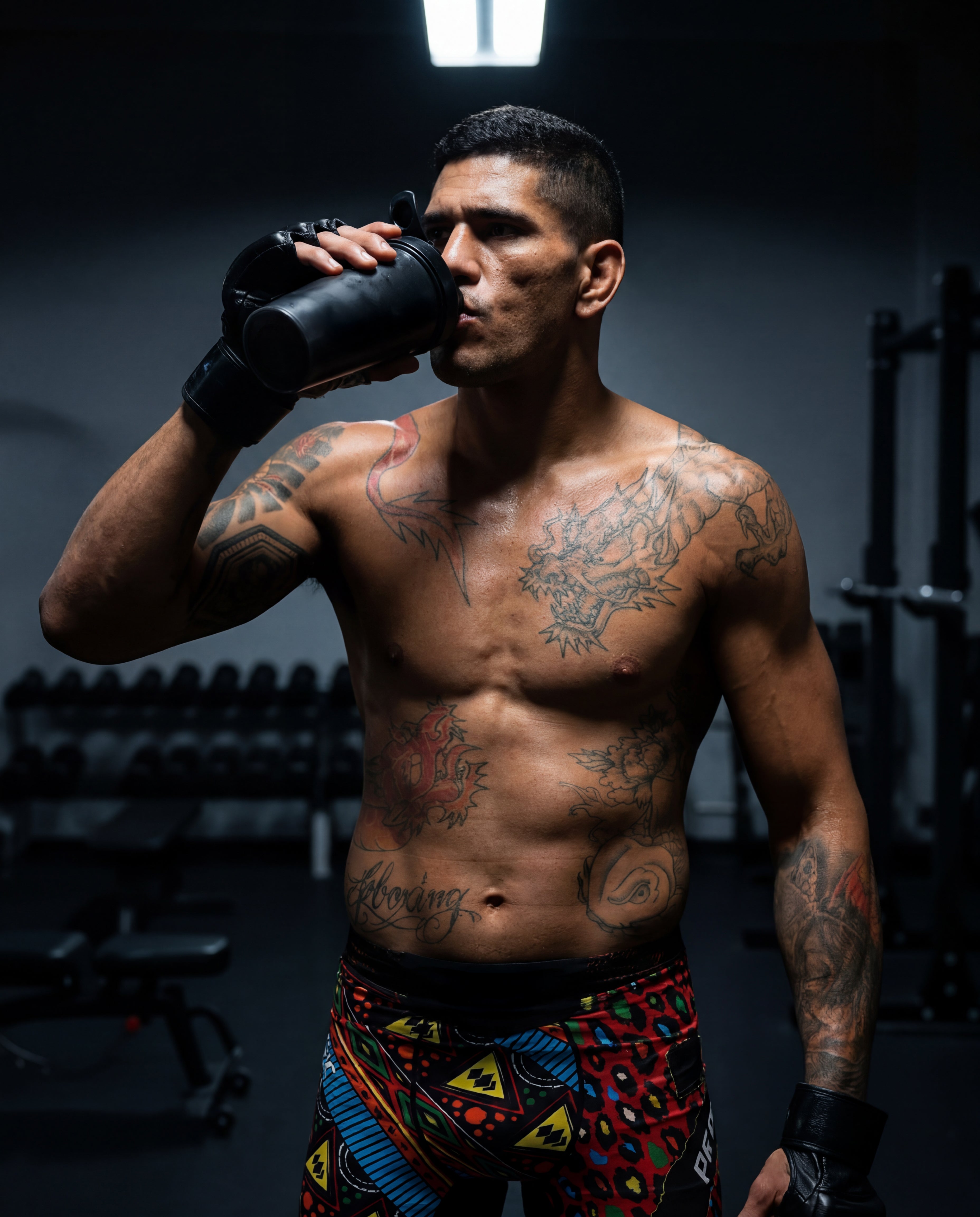 Man with tattoos drinking from a black cup in a gym setting